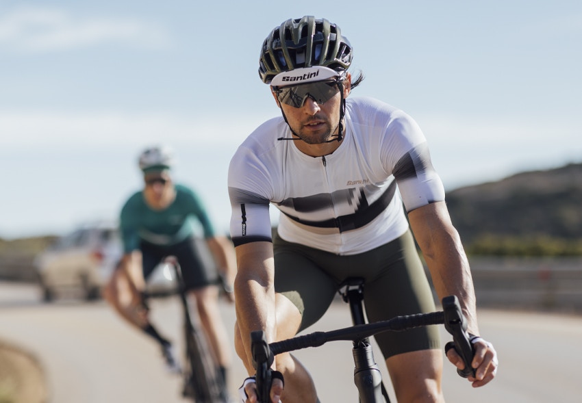 Cyclist wearing a Santini cycling jersey and helmet, riding a bike on a sunny day with another cyclist in the background.